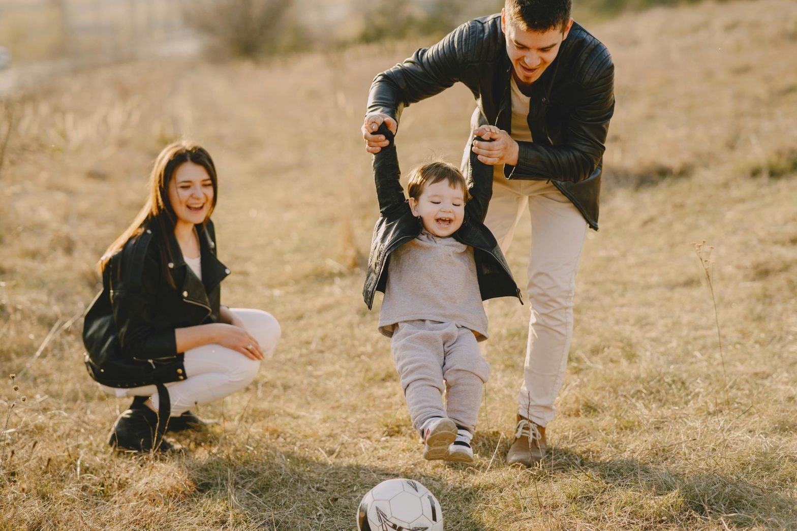 A Happy family of three, where the father is teaching the toddler to walk and the mum is looking at them with pride and joy